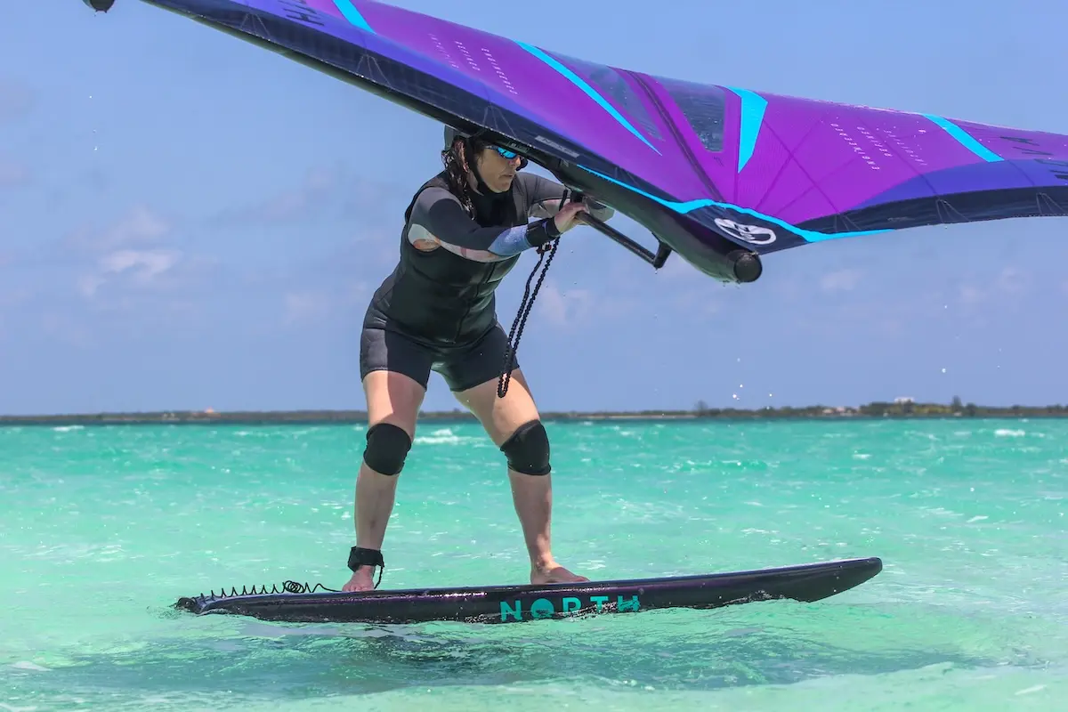 Woman in a wetsuit standing on a North foil board holding a purple wing on crystal clear turquoise water during a women's wingfoil retreat in Belize.