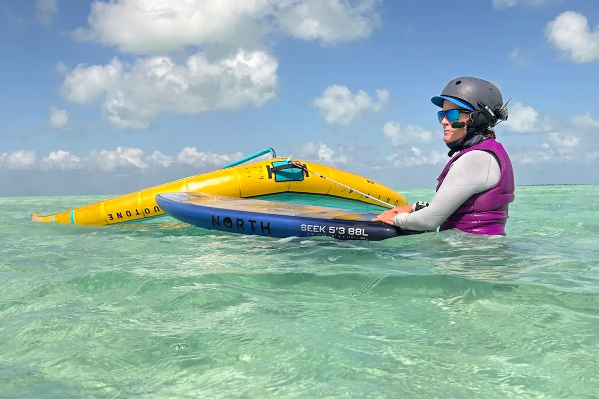 Woman in a purple vest holding a North Seek 5'3 board and a yellow Duotone wing in turquoise shallow Belize water during a women's wingfoil retreat.